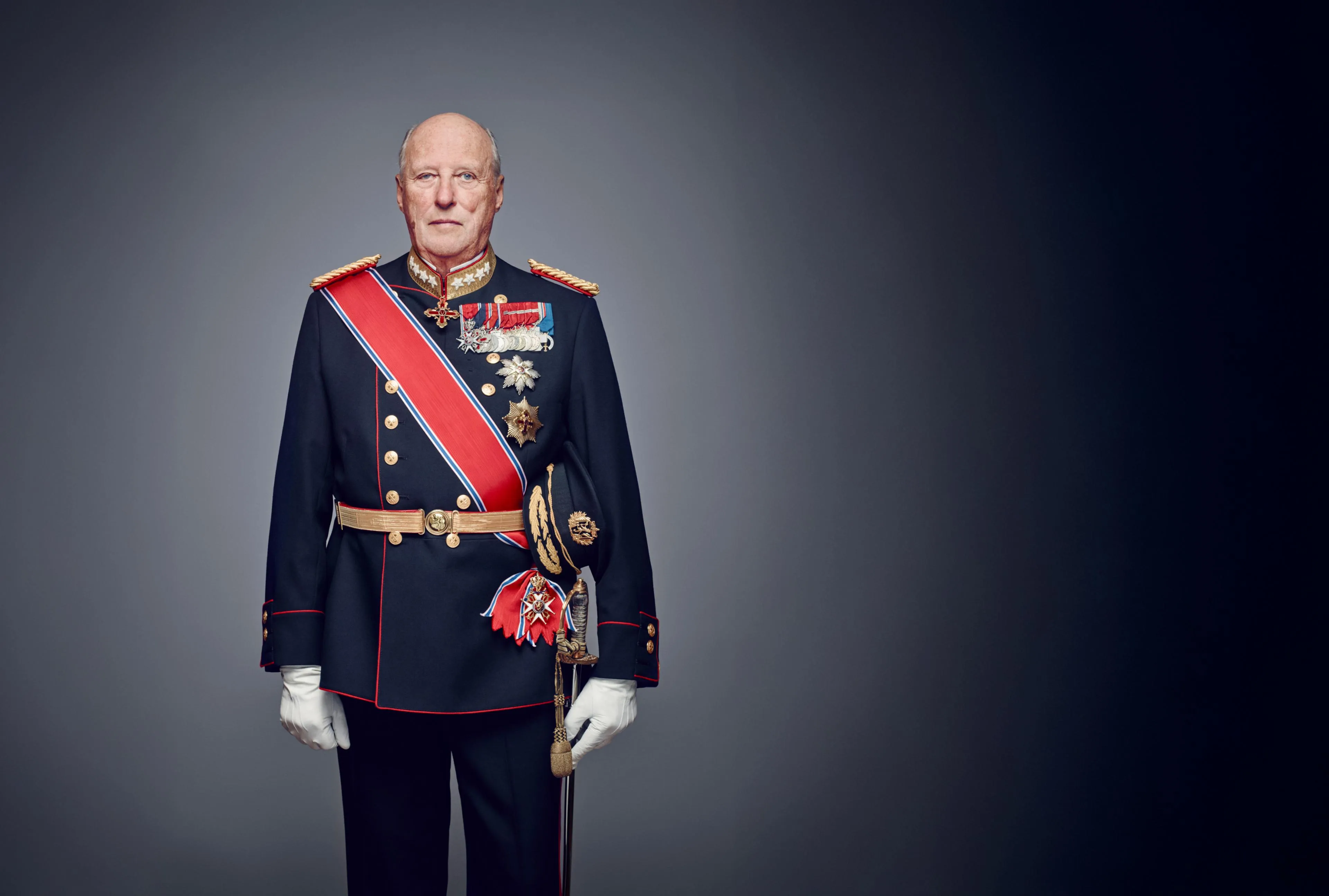 Formal portrait of the late King Harald V of Norway (1937–2026), shown in a dark navy ceremonial military uniform with red trim, a red-white-blue sash, gold epaulettes, medals and decorative orders on the chest, white gloves, and a ceremonial sword, standing against a dark gradient studio background.