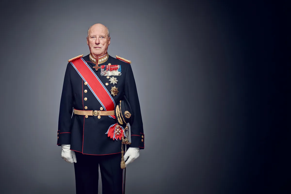 Formal portrait of the late King Harald V of Norway (1937–2026), shown in a dark navy ceremonial military uniform with red trim, a red-white-blue sash, gold epaulettes, medals and decorative orders on the chest, white gloves, and a ceremonial sword, standing against a dark gradient studio background.