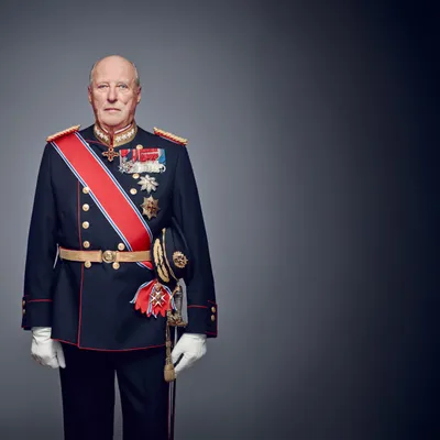 Formal portrait of the late King Harald V of Norway (1937–2026), shown in a dark navy ceremonial military uniform with red trim, a red-white-blue sash, gold epaulettes, medals and decorative orders on the chest, white gloves, and a ceremonial sword, standing against a dark gradient studio background.